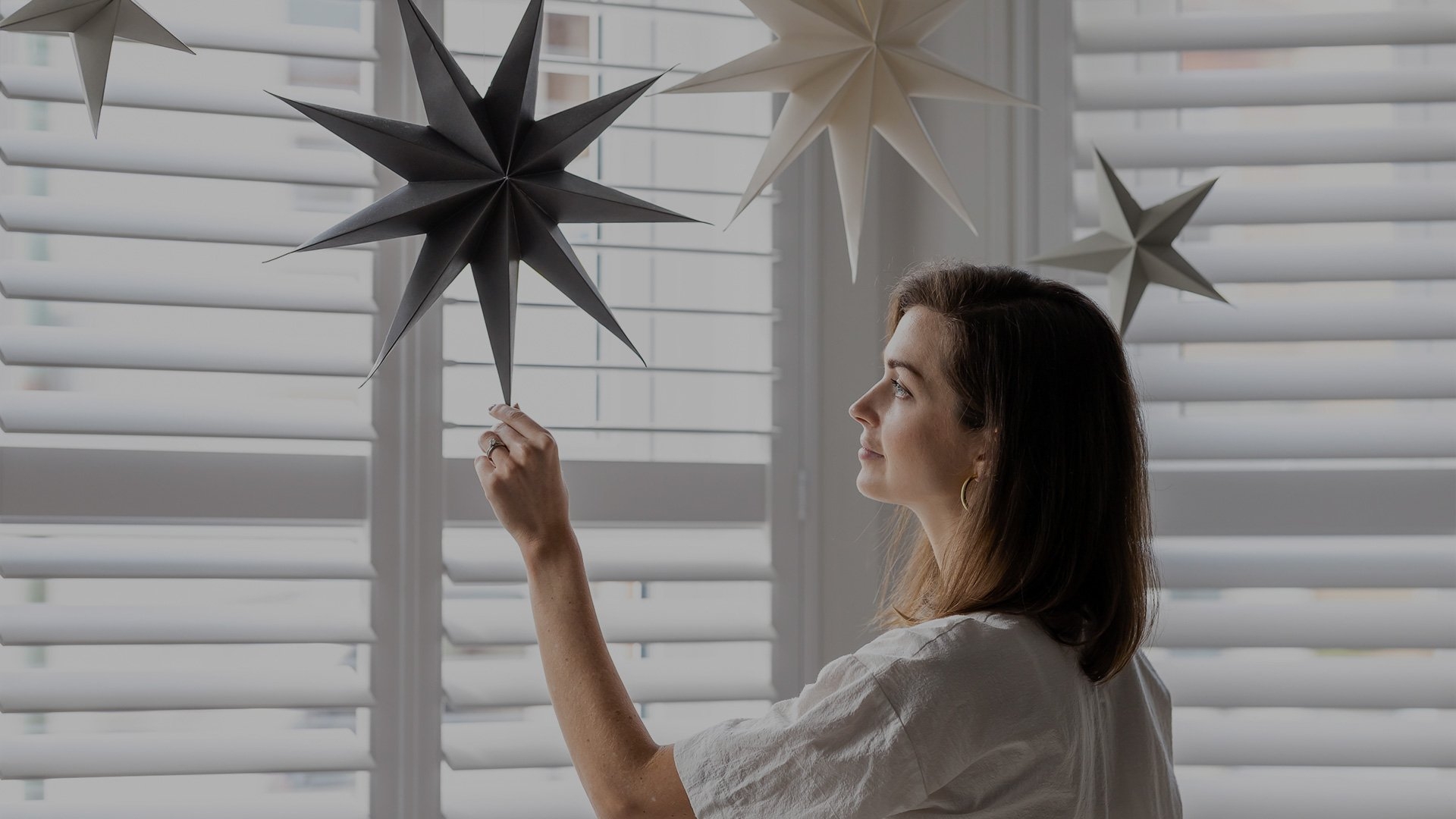 A woman touches a paper star hanging from her ceiling, in front of shuttered windows.